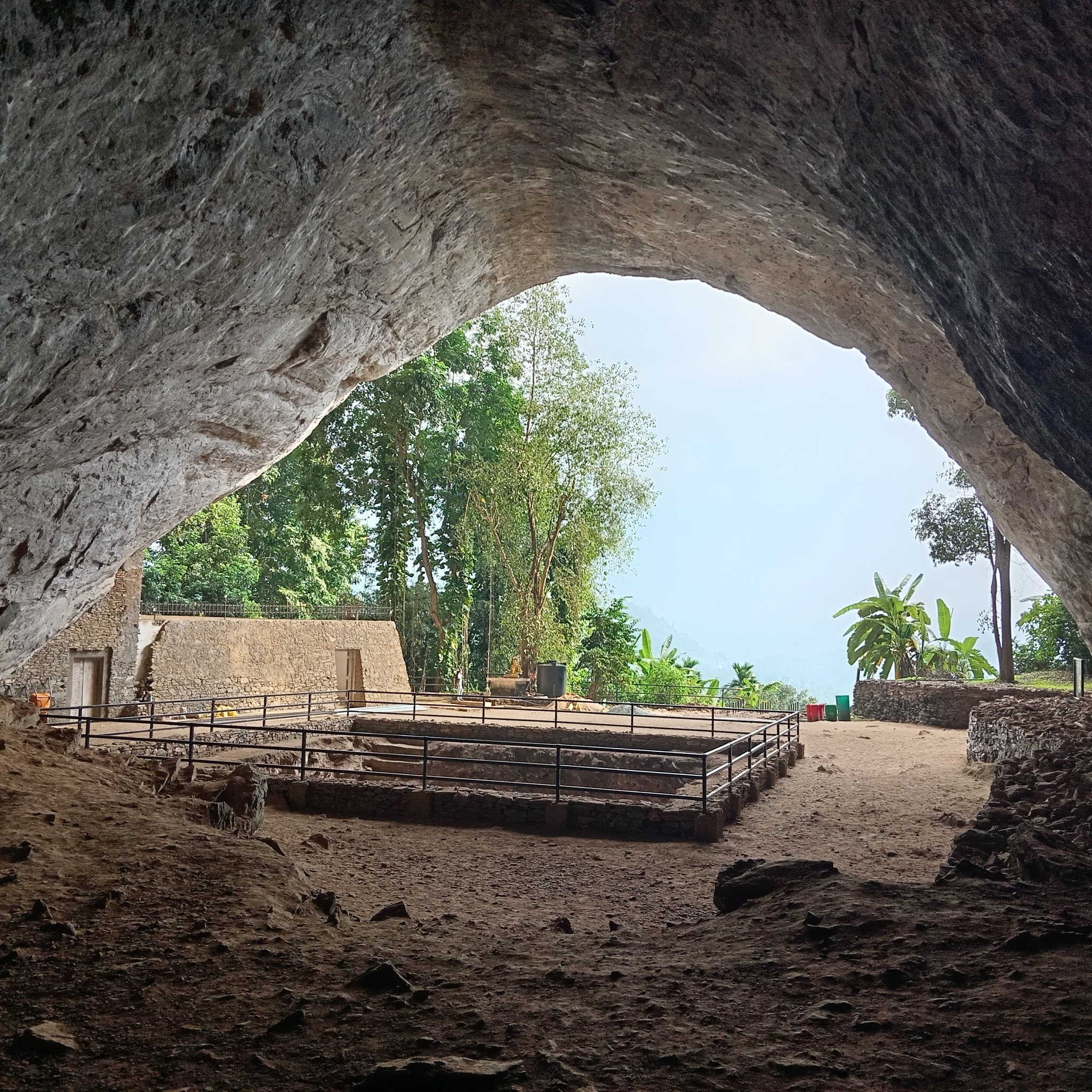 The immense, sunlit entrance of Pahiyangala Cave in Sri Lanka, with a large reclining Buddha statue and lush jungle surroundings.